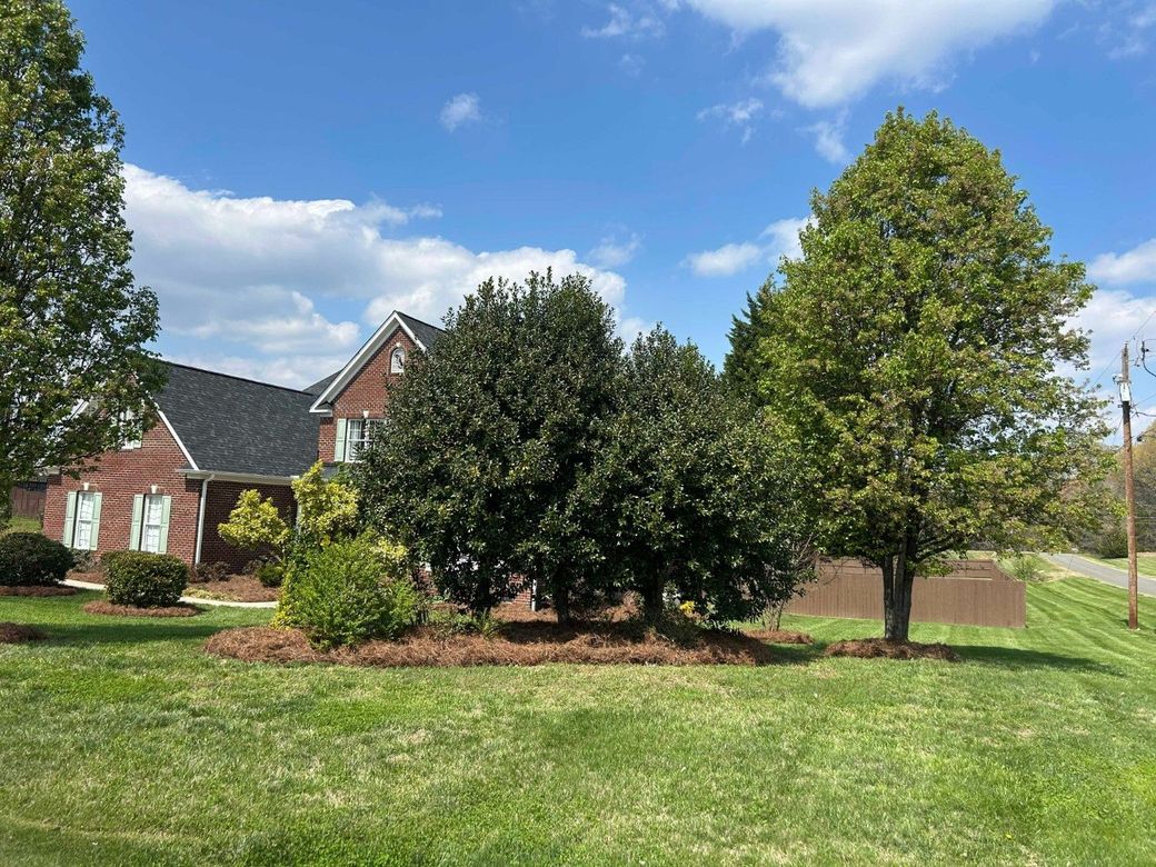 Brick house with large green trees and a grassy front yard under a blue sky