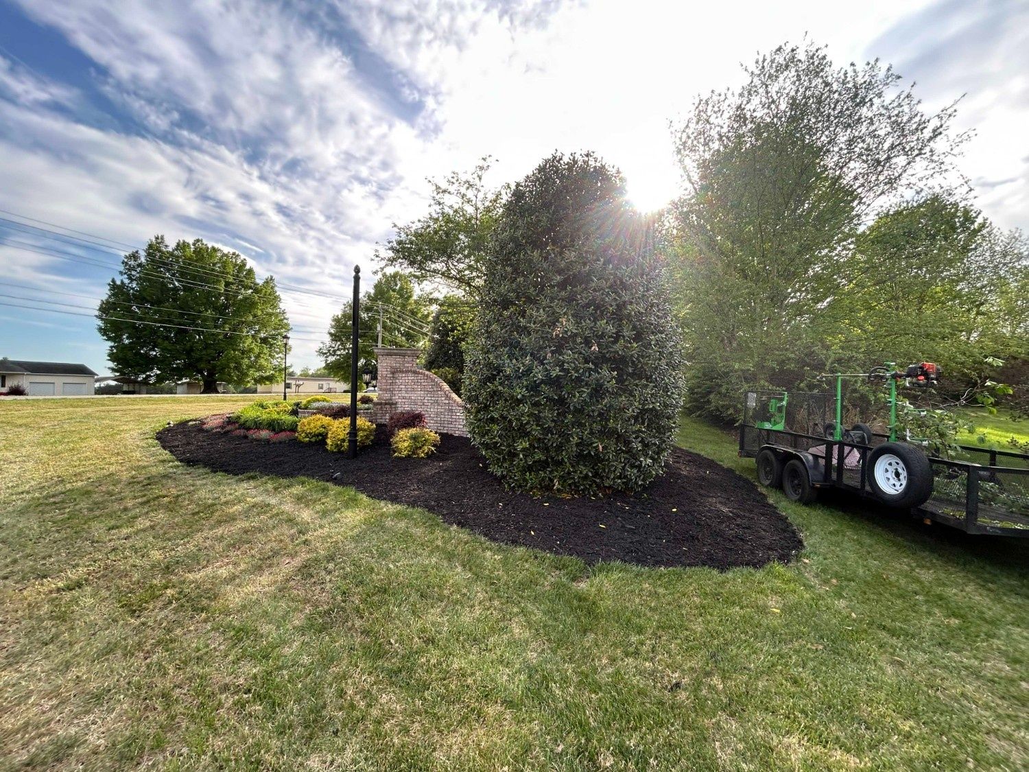 Sunny yard with trimmed shrubs, mulched flower beds, and a utility trailer on the right