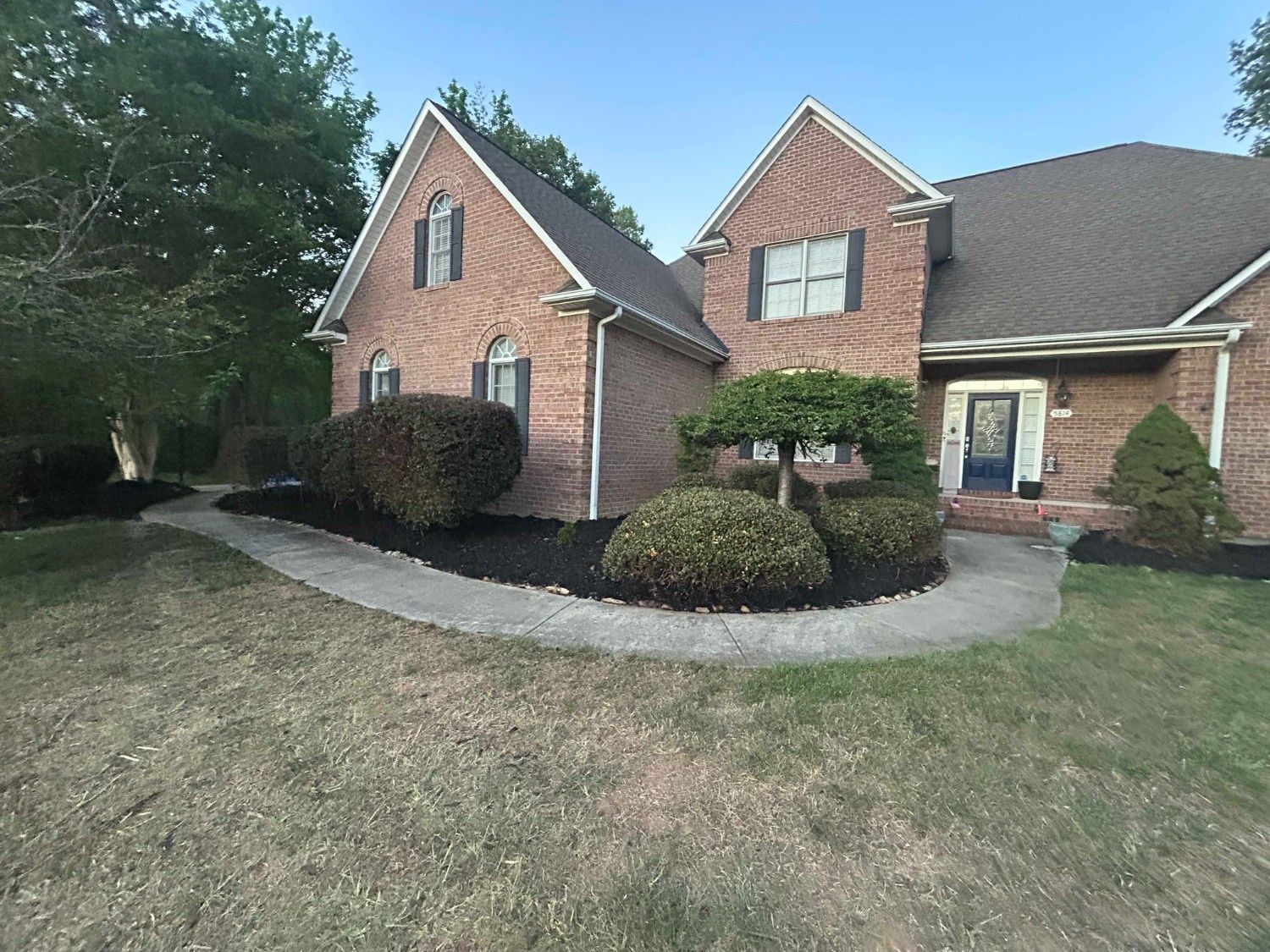 Brick suburban house with gabled roof, shrubs, and a curved concrete walkway in front of a lawn