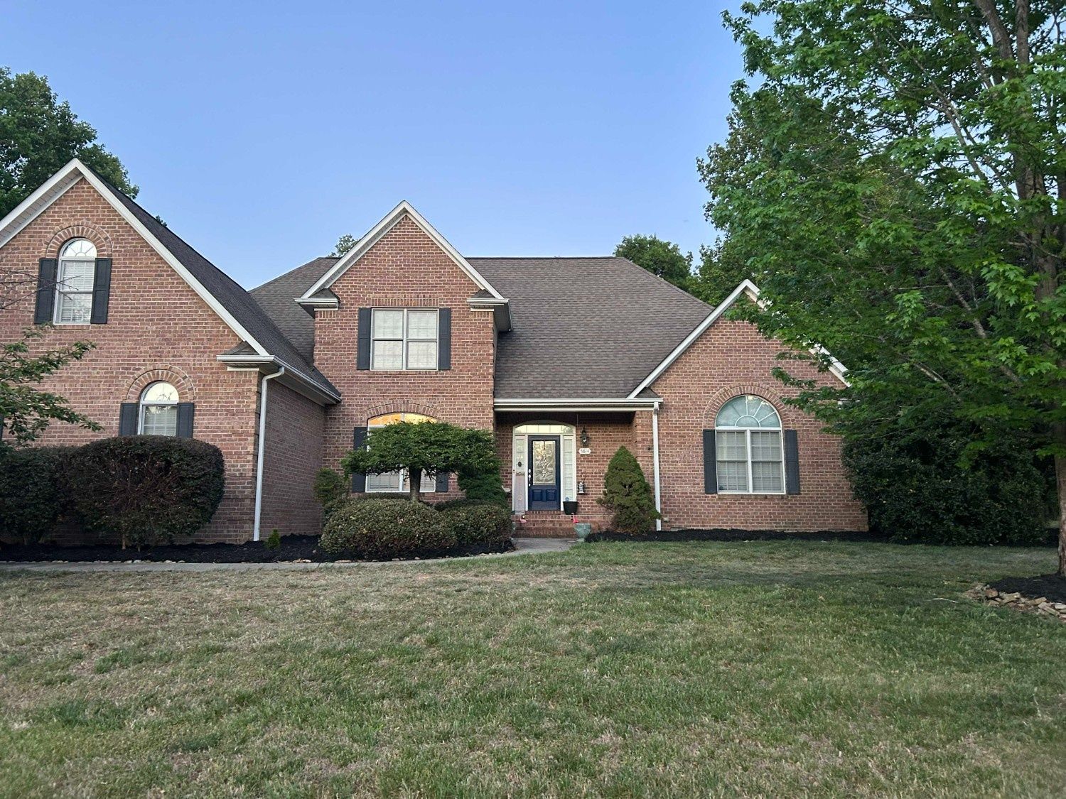 Brick suburban house with a front lawn, trees, and a driveway under a clear blue sky