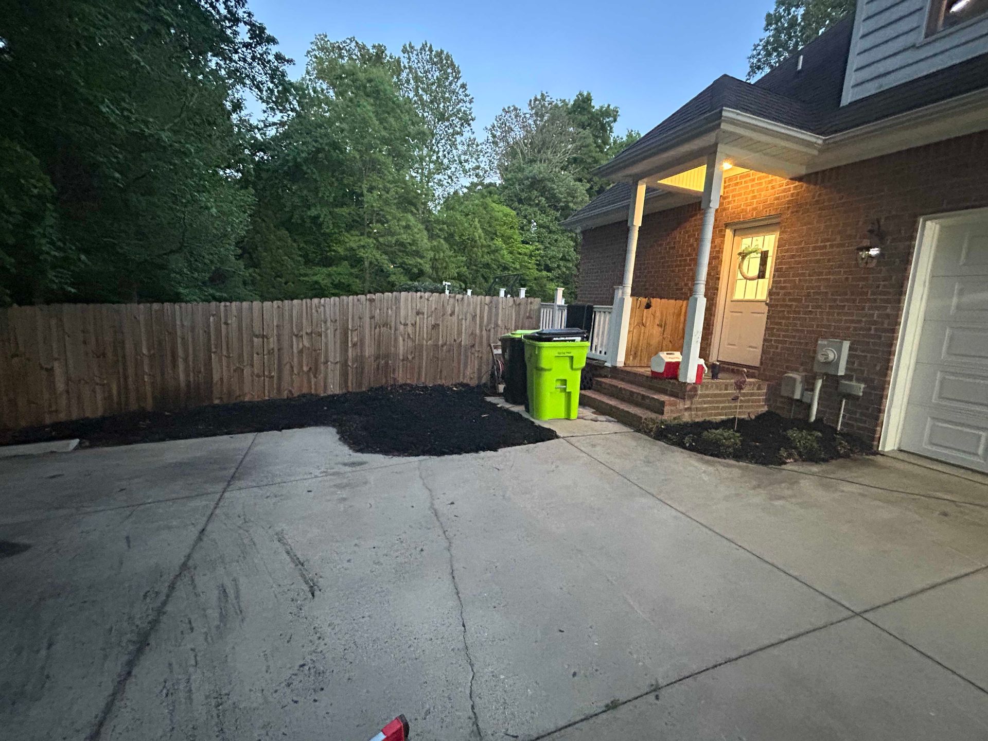 Driveway beside a brick house with a bright green trash bin, wooden fence, and trees in the background