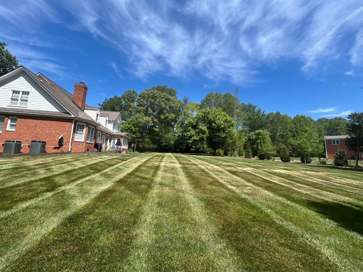 Large mowed lawn with striped grass, red-brick house on the left, trees, and blue sky with clouds