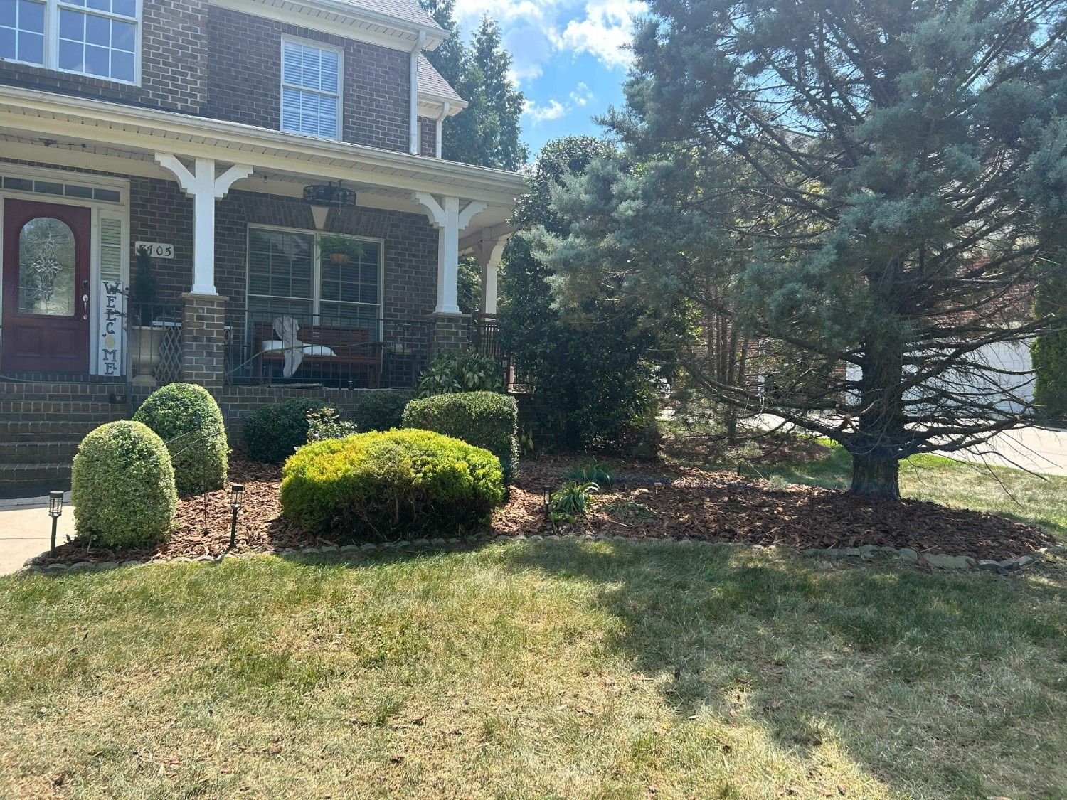 Suburban brick house with porch, trimmed shrubs, and a sunlit front lawn under trees.