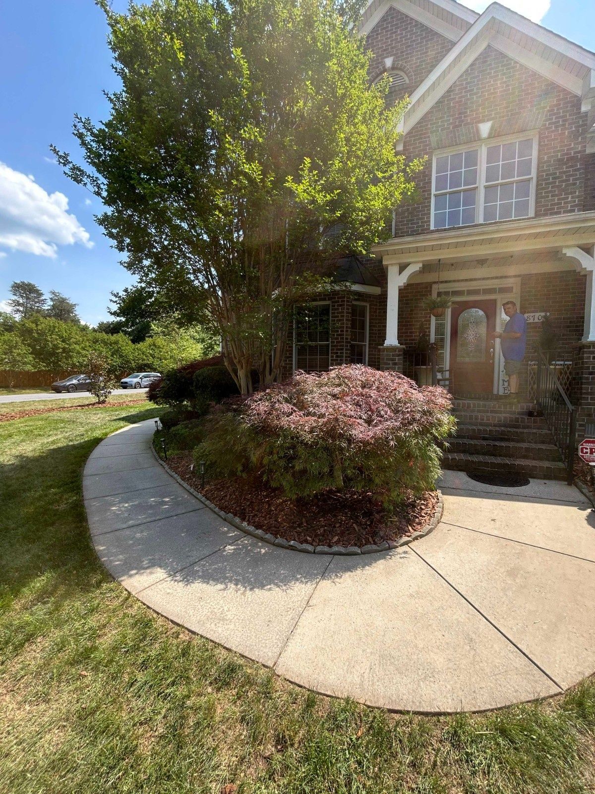 Brick house entrance with curved sidewalk, porch columns, and landscaped bushes under a sunny sky
