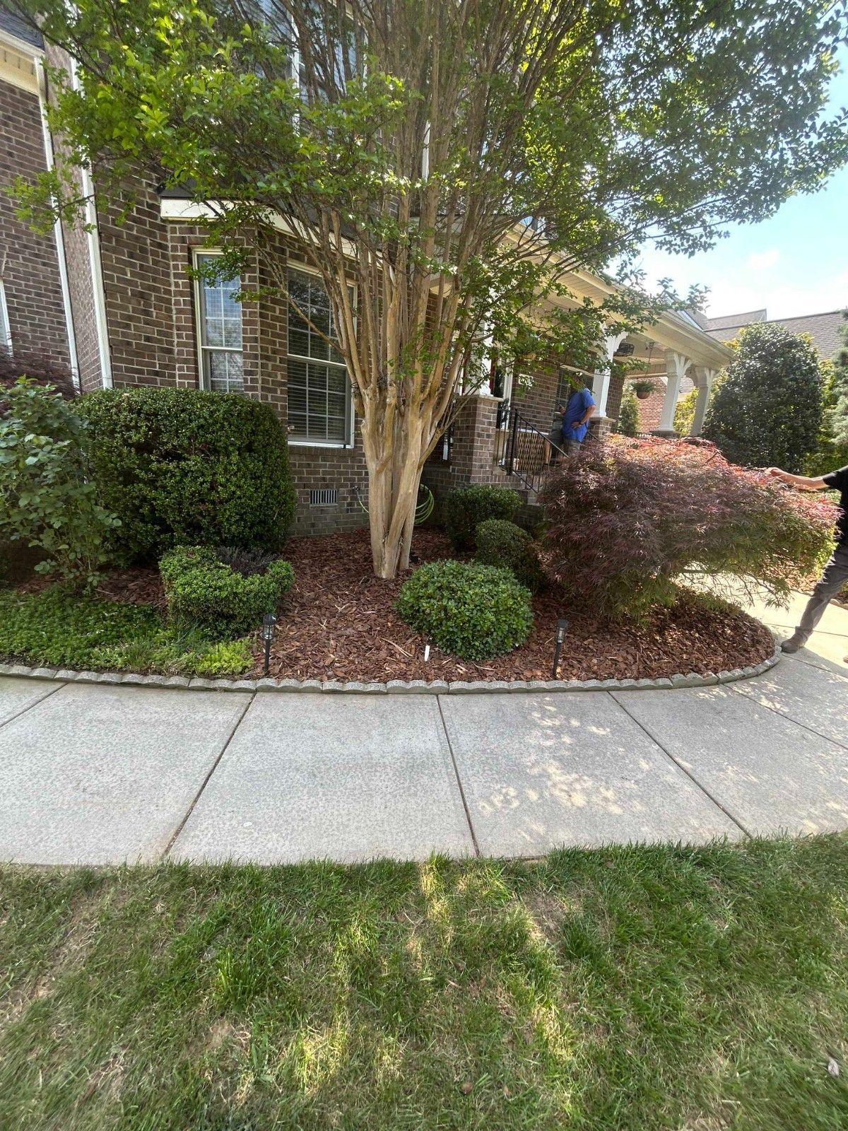 Landscaped brick house front with sidewalk, trees, shrubs, and a person near the porch