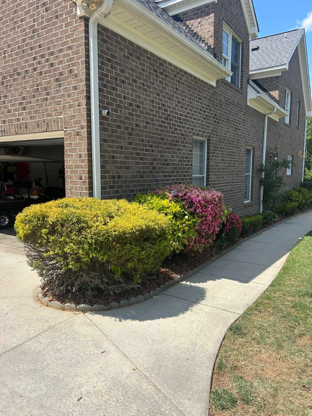 Brick house exterior with garage, curved driveway, and trimmed green and pink shrubs beside the sidewalk