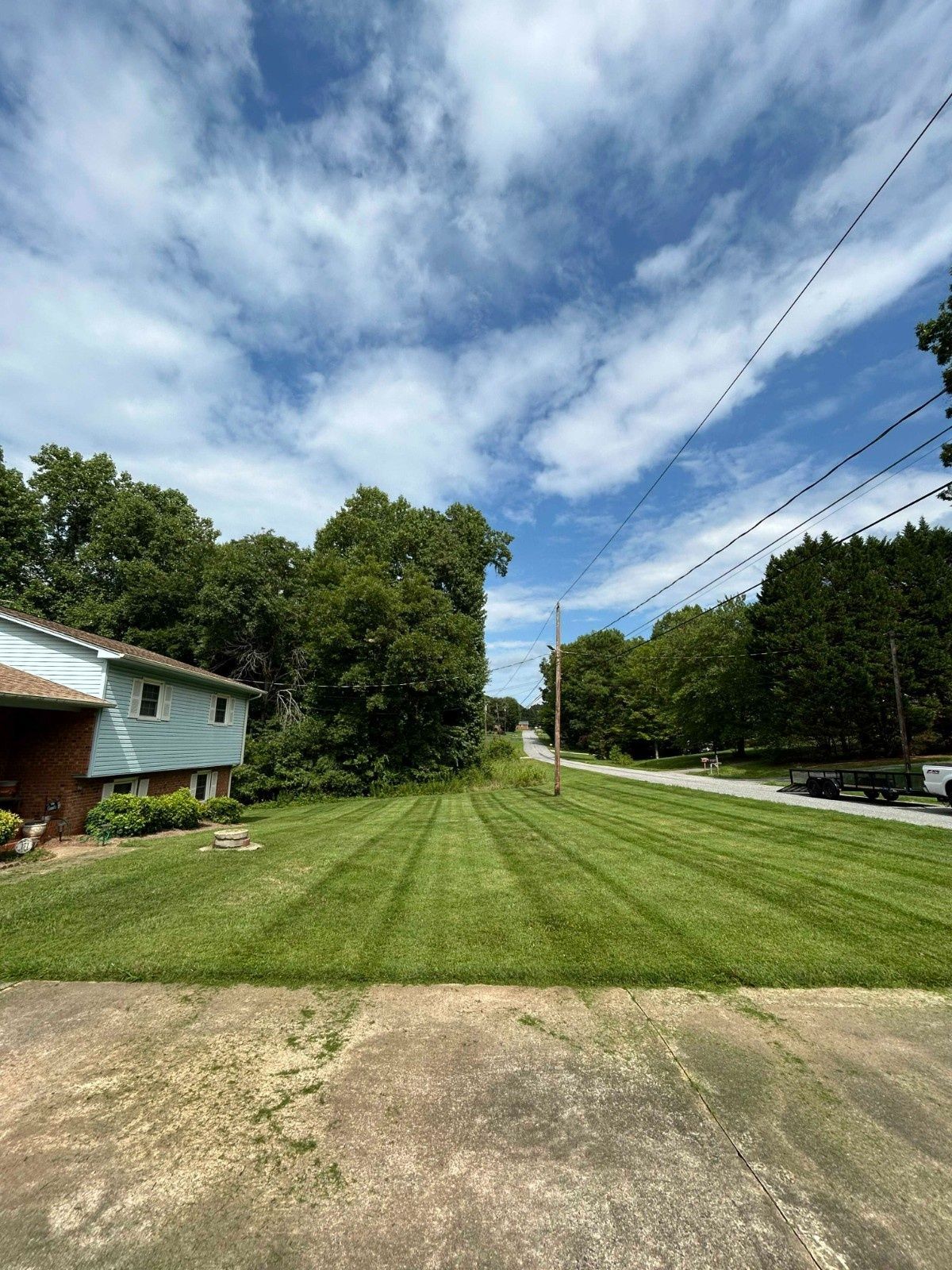 Residential lot with mowed grass, a concrete driveway, trees, and power lines under a partly cloudy sky