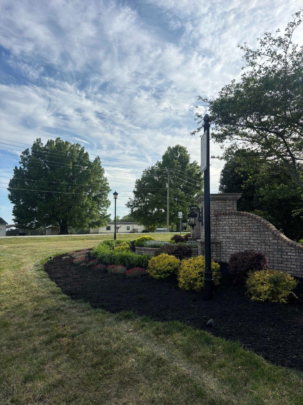 Landscaped grassy hill with shrubs, a stone wall, and tall trees under a cloudy sky
