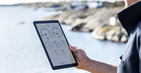 A person holding a tablet displaying digital gauges while standing outdoors near a body of water and rocky shoreline.