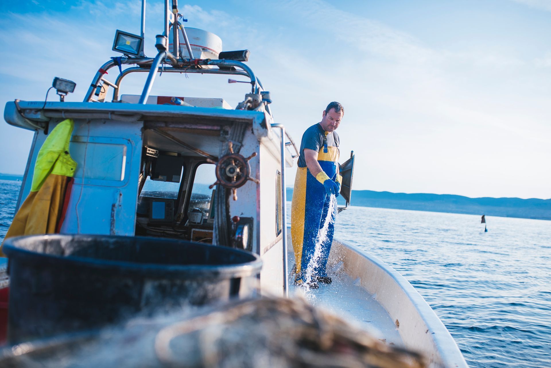 Fisherman on boat rinsing deck using marine hoses during open sea fishing trip. A person in waterproof work gear stands on the deck of a boat, casting a fishing net into the bright, open sea.