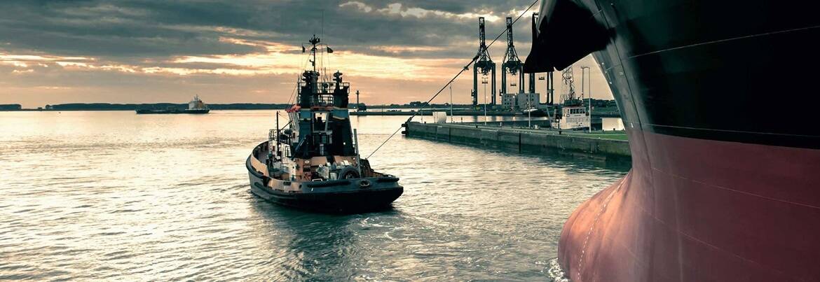 A tugboat maneuvering near the bow of a large ship in a harbor at sunset, with port cranes visible in the background.