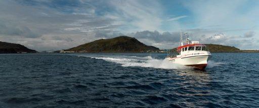 A white and red boat moves quickly across calm blue water toward an island under a cloudy sky.