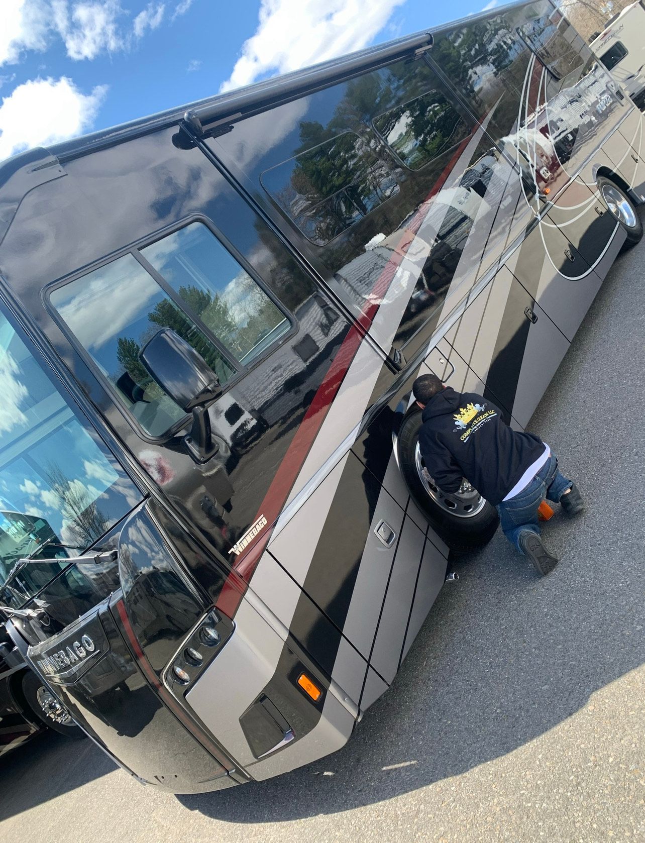Mechanic working on the tire of a large black and gray RV on a paved lot.