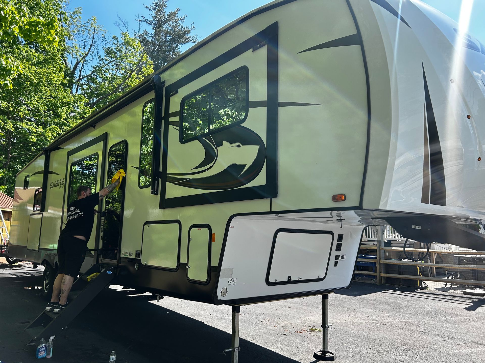 Person cleaning a large RV, which is parked outdoors in a sunny setting. The RV is beige, black, and white.