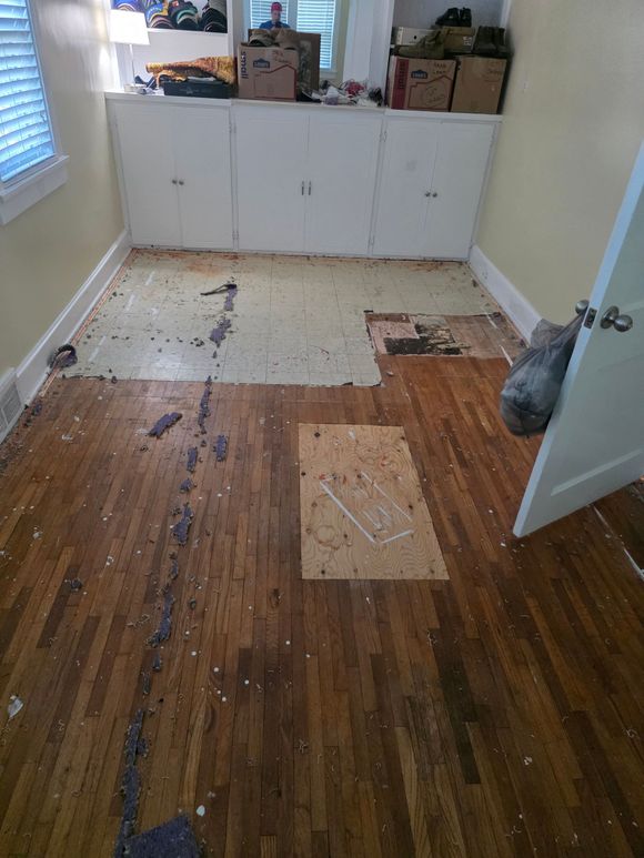 Damaged wooden floor with missing panels and exposed underlayment in a small room with white cabinets.