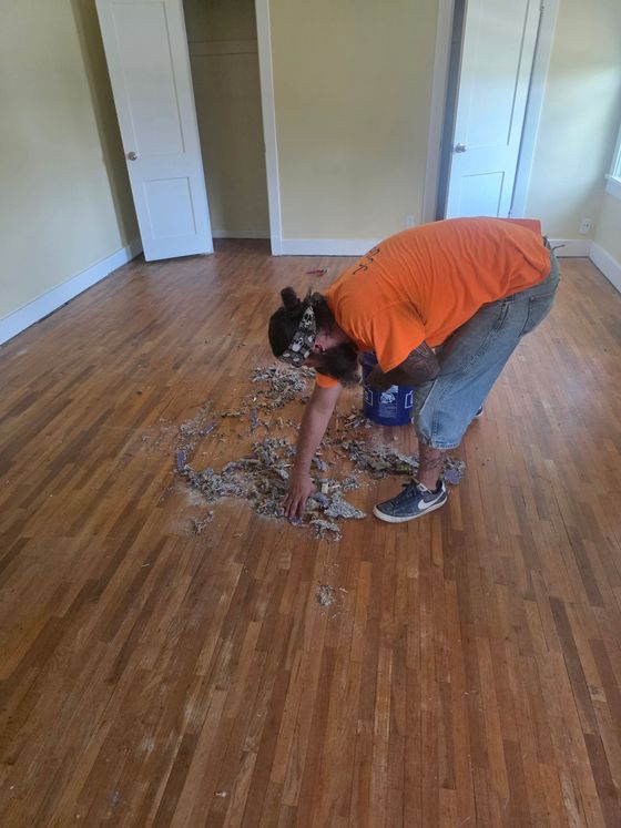 Person cleaning a hardwood floor with a rag in an empty room, wearing an orange shirt and jeans