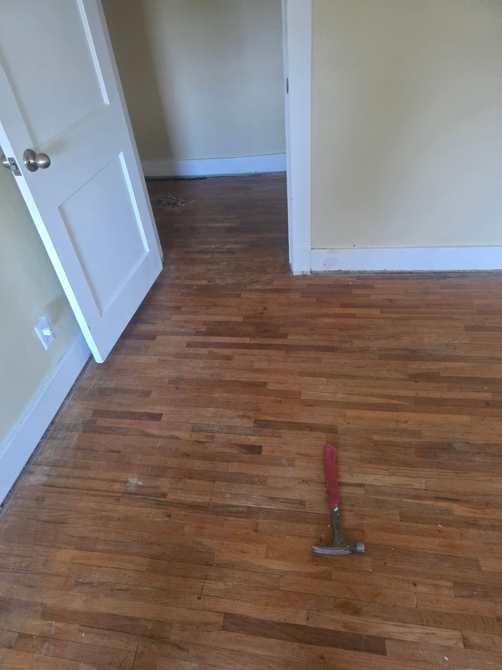 Empty room with wooden floor, open white door, and a hammer on the floor.