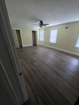 Empty living room with dark wood floors, white walls, ceiling fan, and windows with blinds.