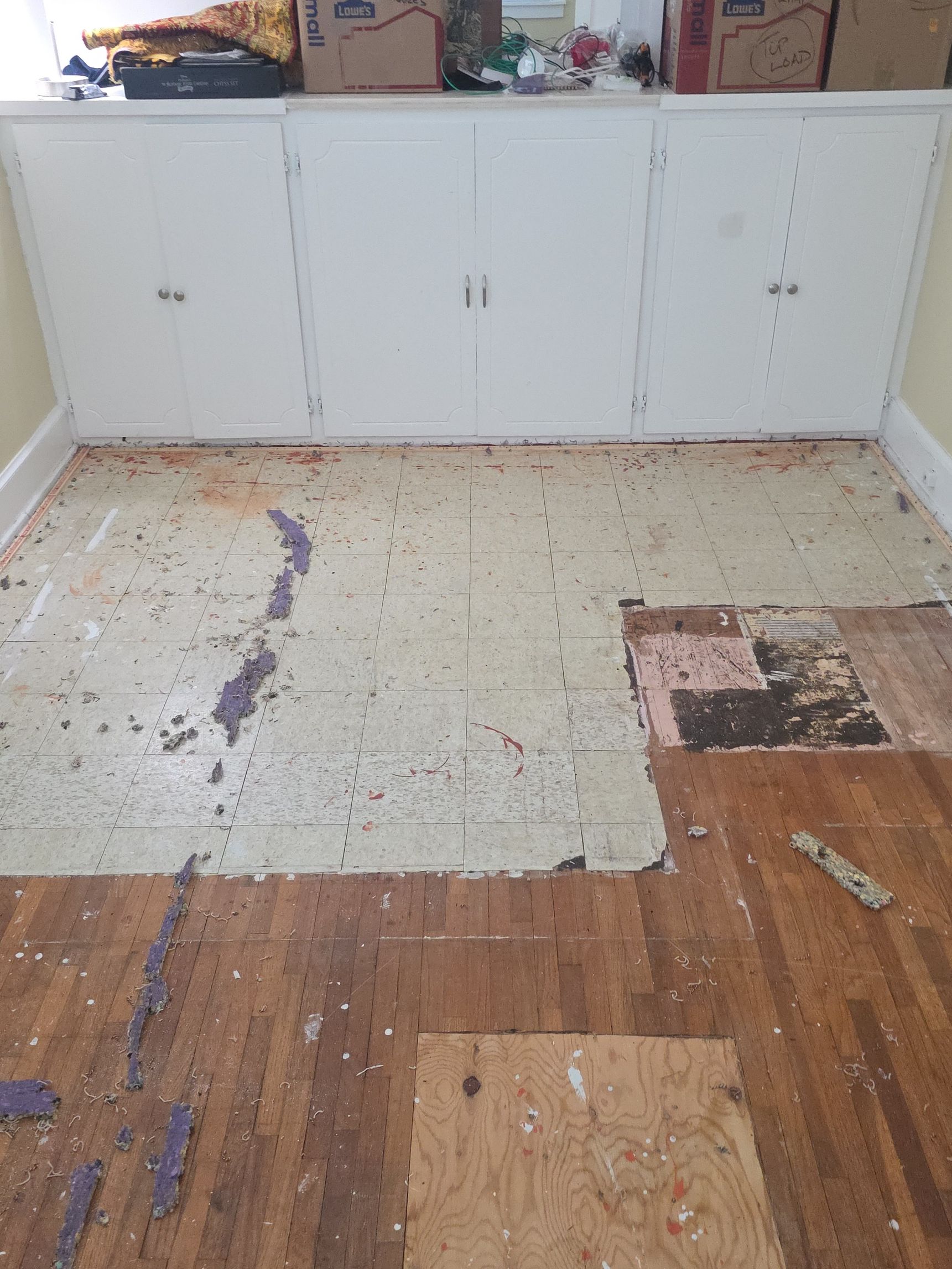 Damaged kitchen floor with peeling linoleum and exposed wood in front of white cabinets.