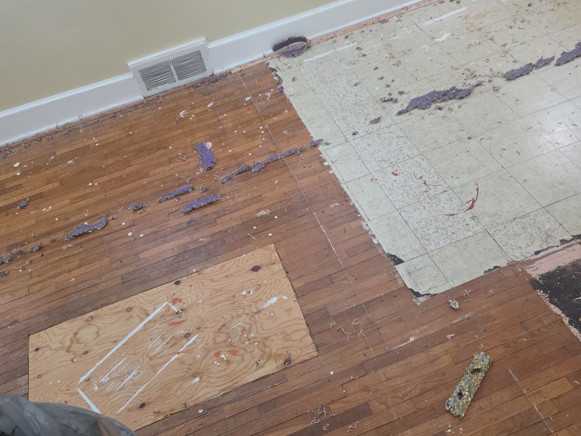 Damaged wood and tile flooring with missing patches near a wall vent
