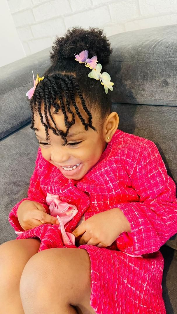 Young Black girl in pink robe smiles while looking down. Her hair is styled with small braids and flower clips.