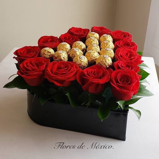 Ramo de rosas rojas y chocolates dorados en una caja negra sobre una superficie blanca.