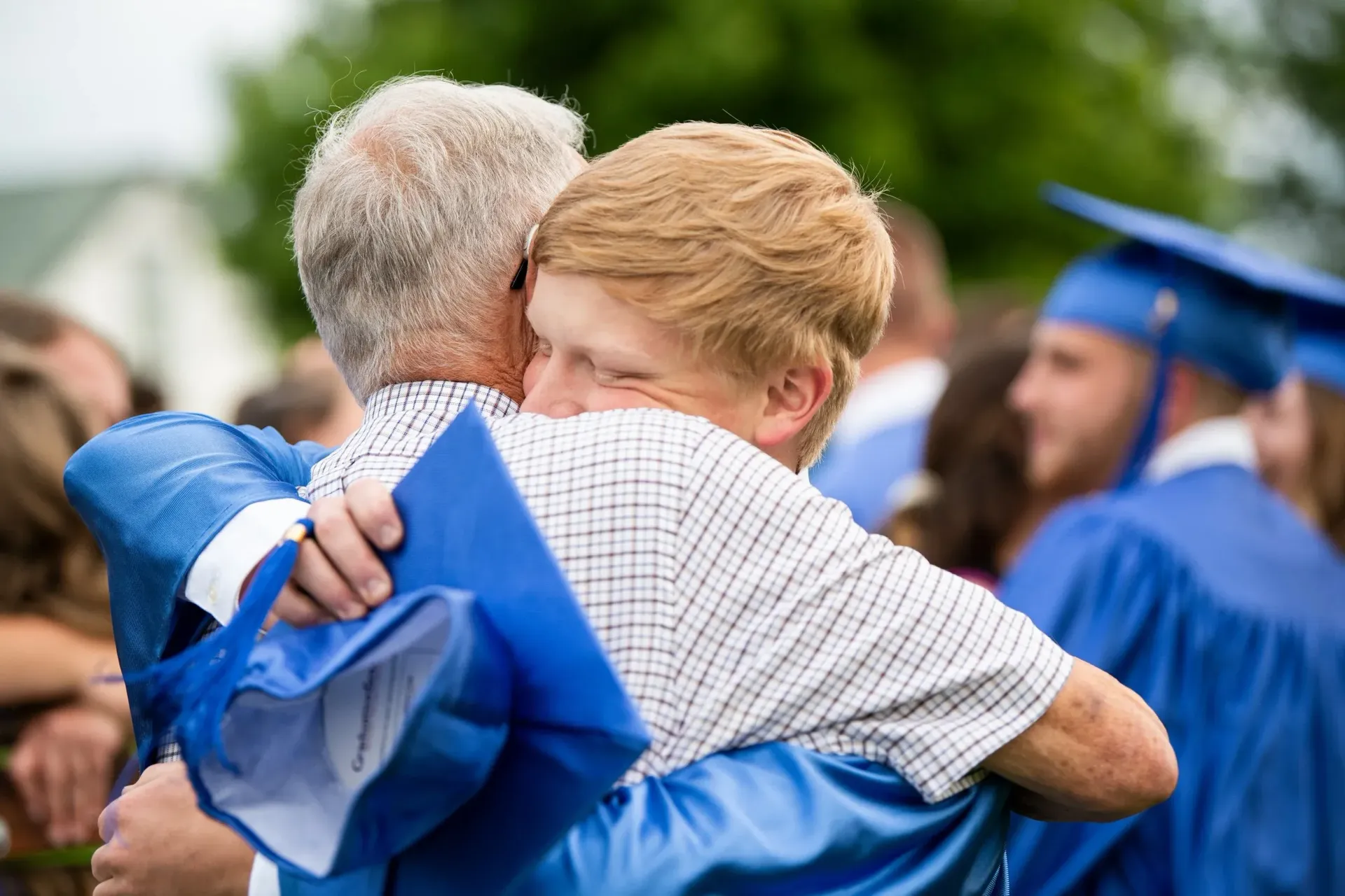 Man in graduation gown hugs another man; outdoors, blurred background.