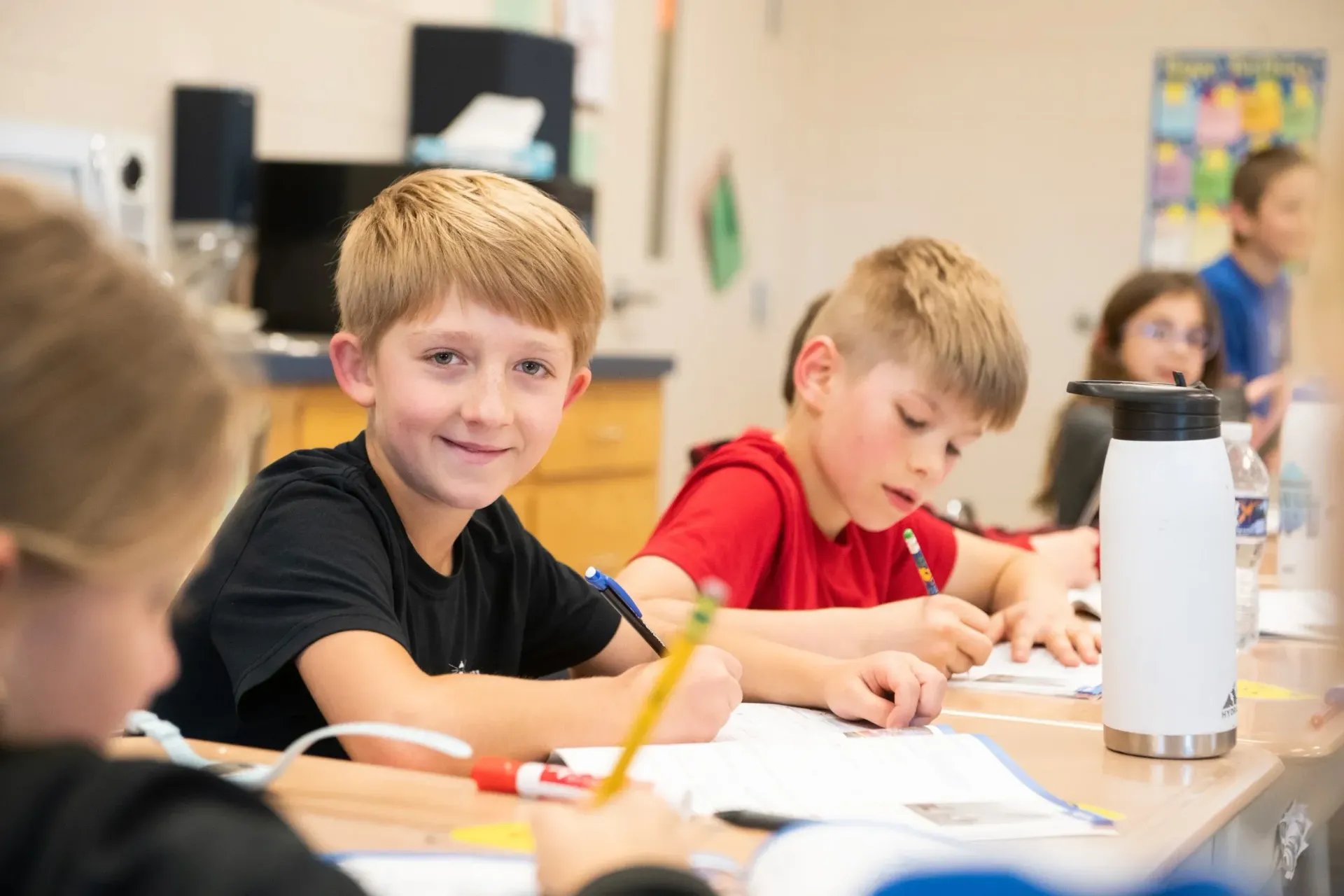 Boy smiles at the camera while writing at a desk with other children in a classroom.