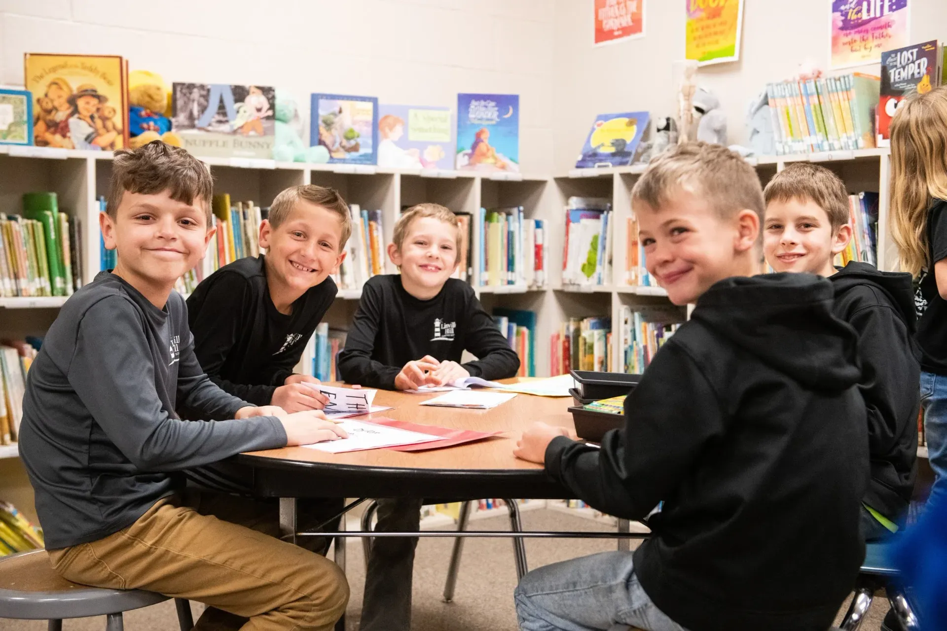 Four smiling children sit at a round library table working on papers with bookshelves in the background.