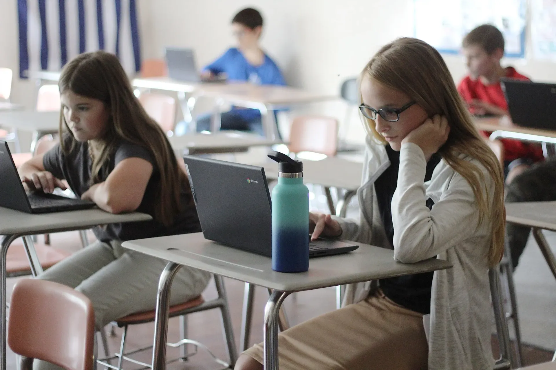 Students seated at desks in a classroom, working on laptops with a water bottle on a desk in the foreground.