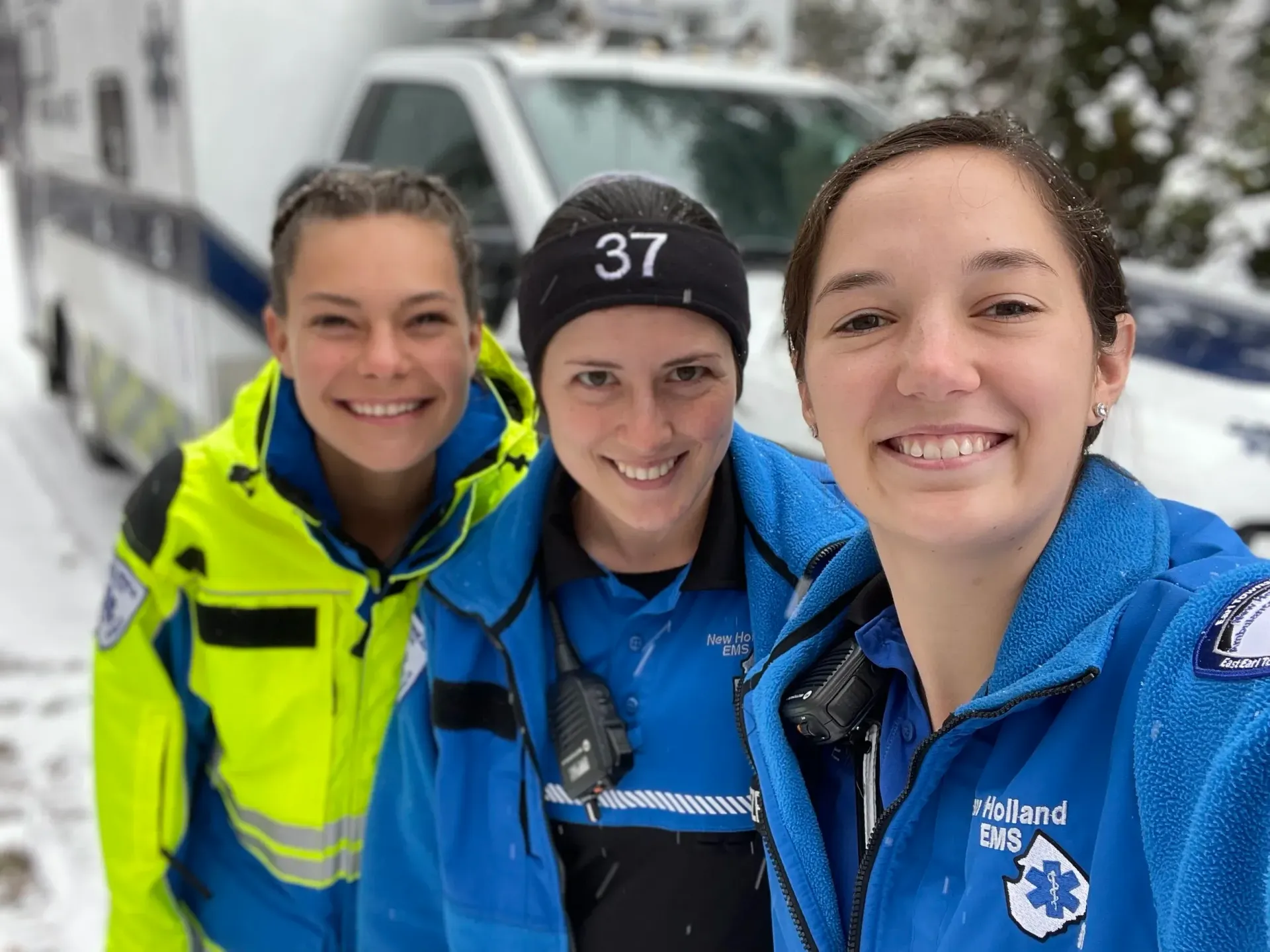 Three emergency medical workers smile in the snow in front of an ambulance.