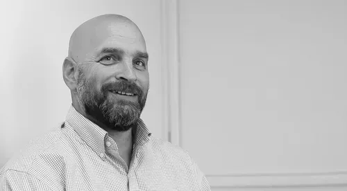 Bald man with a beard, smiling, wearing a light-colored button-down shirt, set against a plain background.
