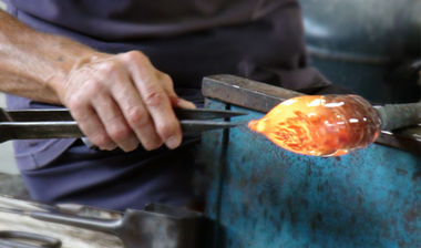 Un soplador de vidrio da forma al vidrio fundido incandescente con unas pinzas sobre un horno azul.