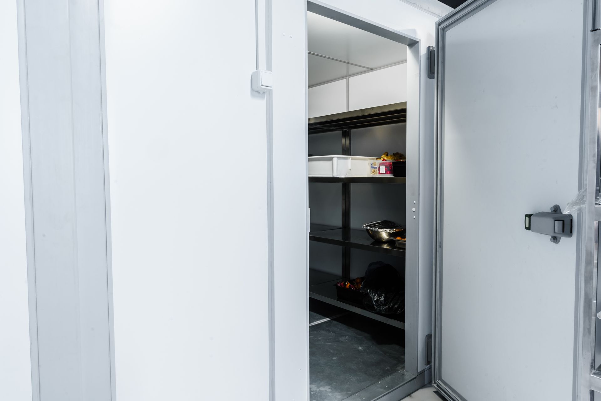 Open white walk-in cooler with metal shelves and a partly open door Open white walk-in cooler with metal shelves and a partly open door