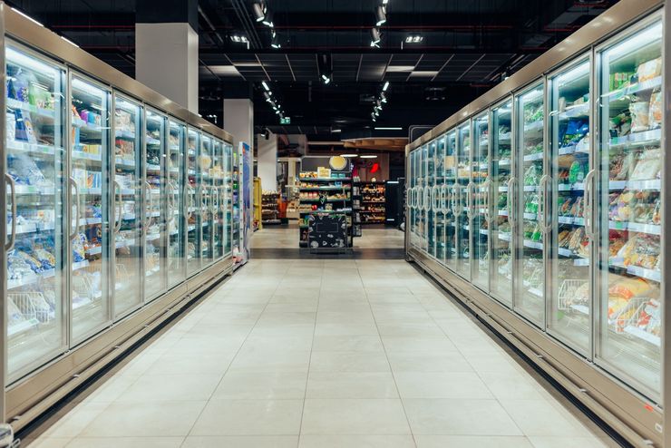 A supermarket freezer aisle with glass doors and bright overhead lights leading to shelves at the back. A supermarket freezer aisle with glass doors and bright overhead lights leading to shelves at the back.