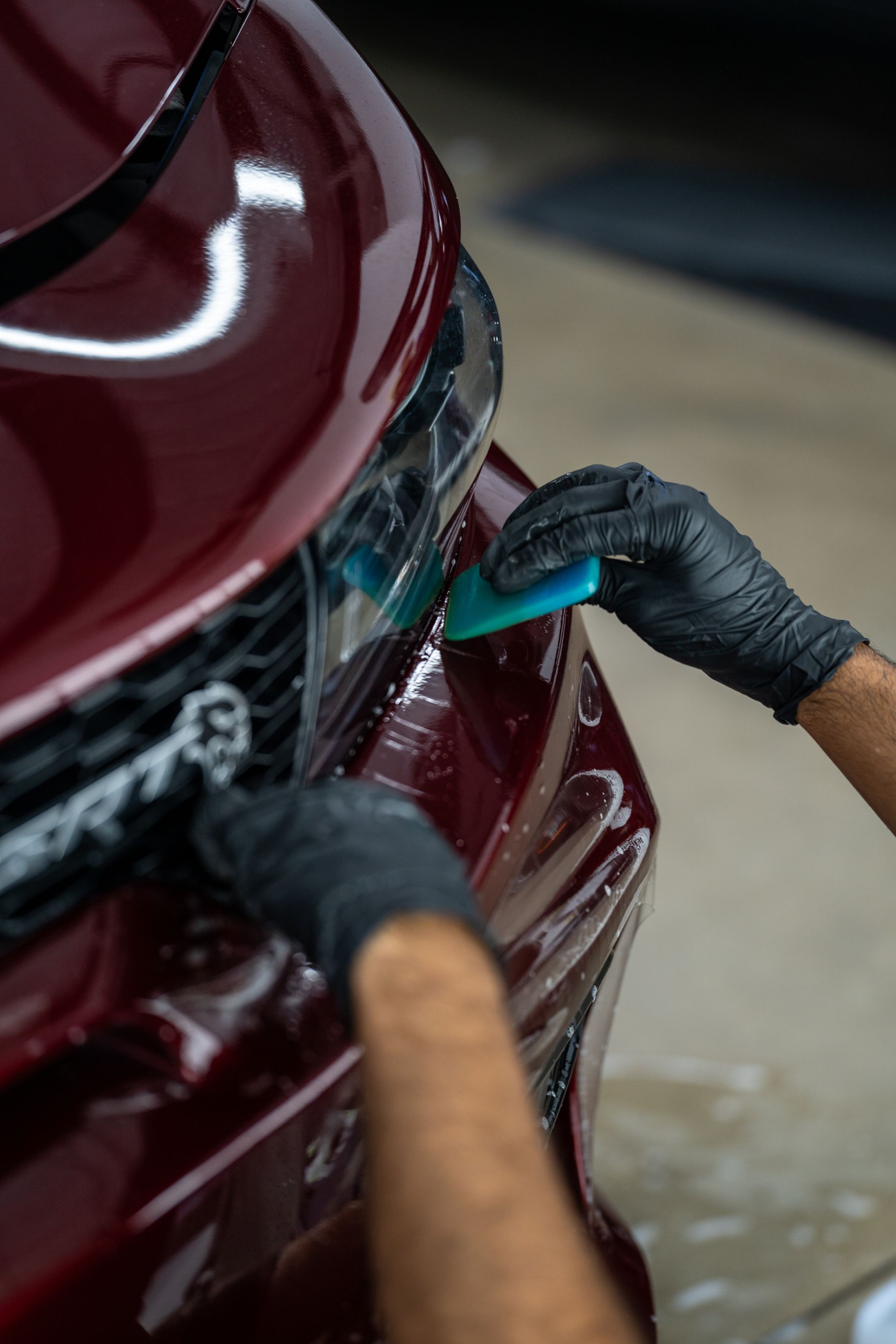 Person applying protective film to a red car's bumper, using a blue squeegee, wearing black gloves.