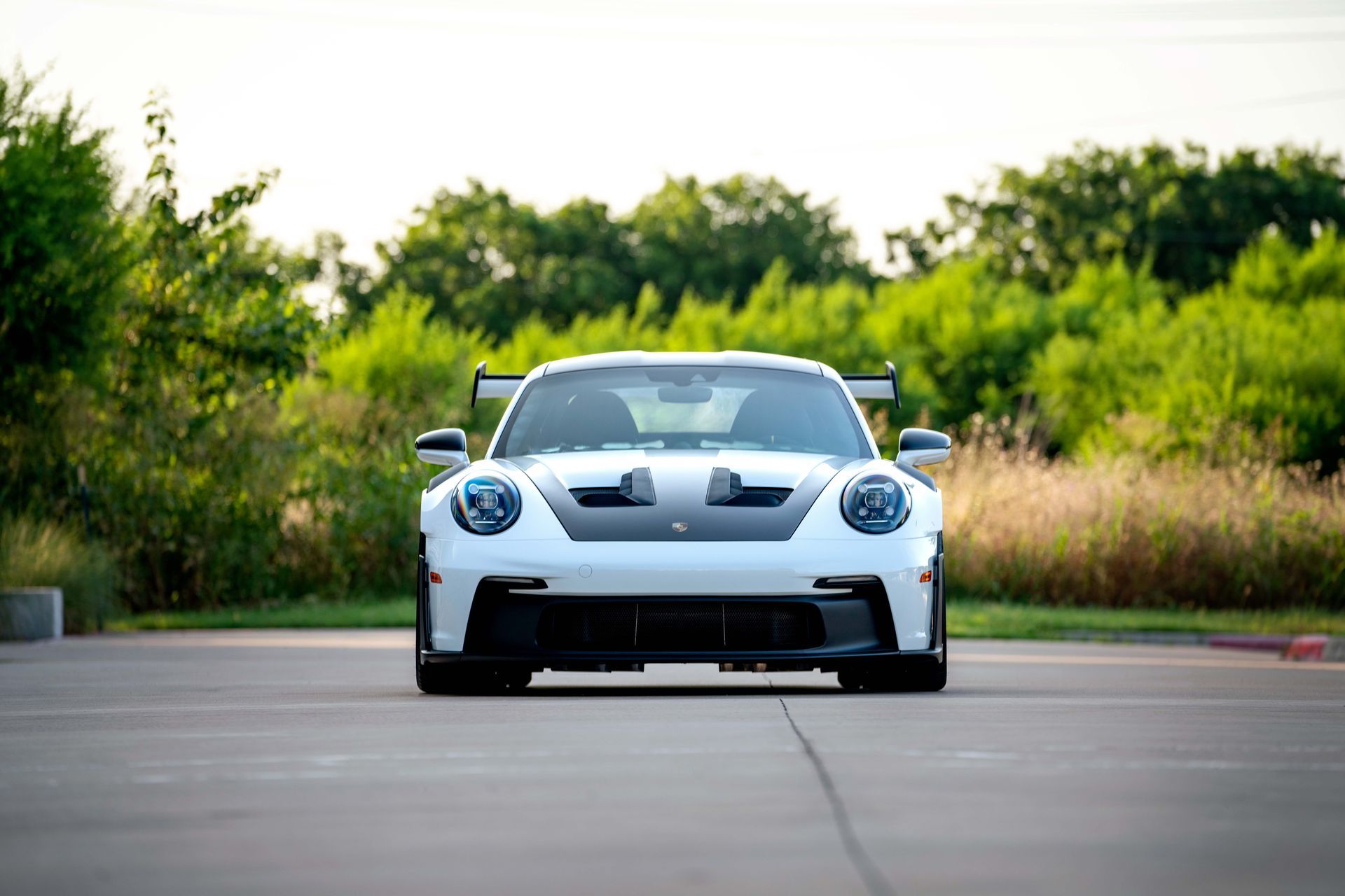 White Porsche 911 GT3 RS sports car parked on a concrete surface, front view. Black accents, trees in the background.