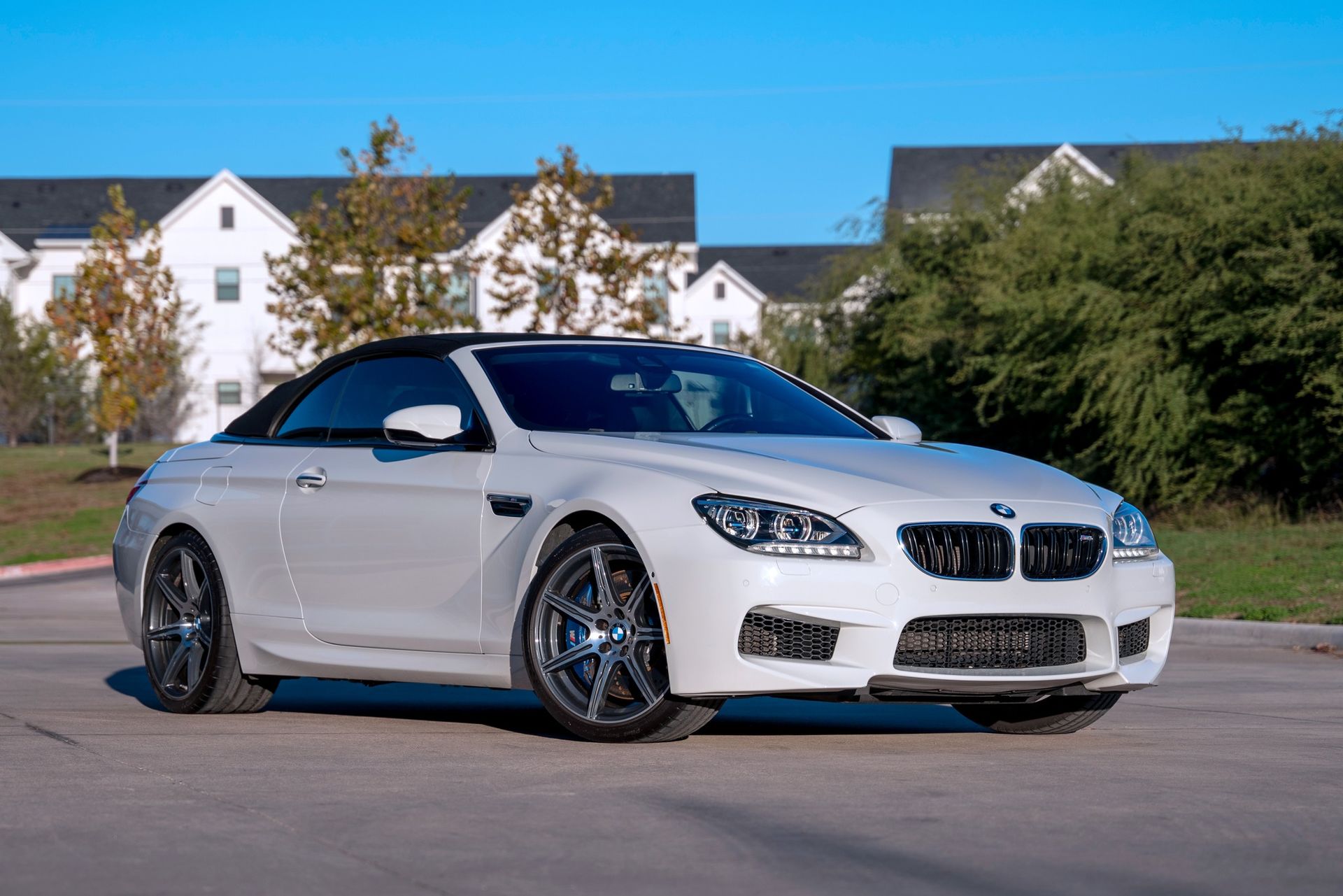 White BMW convertible parked on a paved area, with apartment buildings and trees in the background.