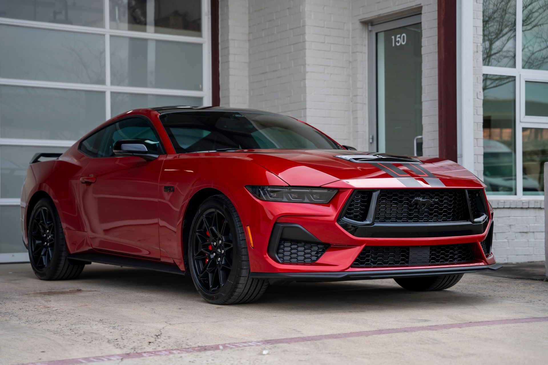 Red Ford Mustang coupe parked in front of a building with black wheels and racing stripes.