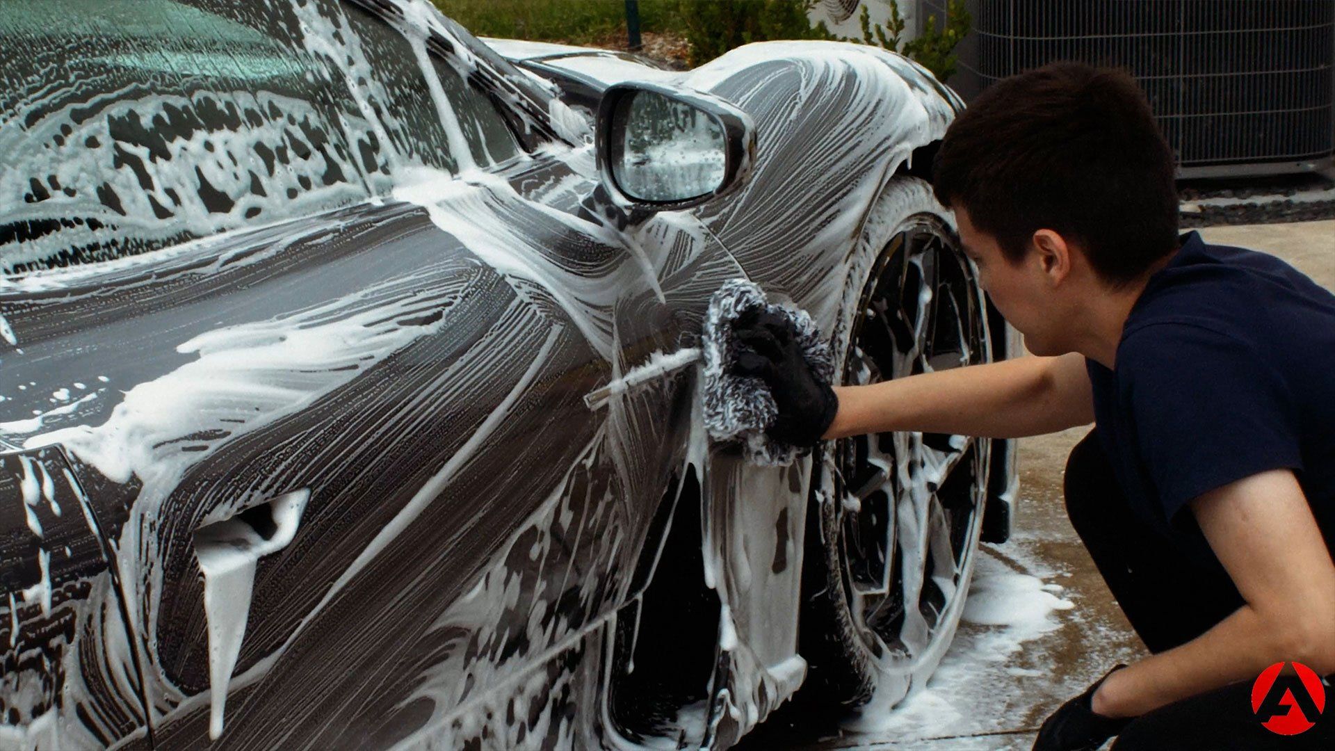 Person washing a dark gray sports car with soapy water using a black mitt.