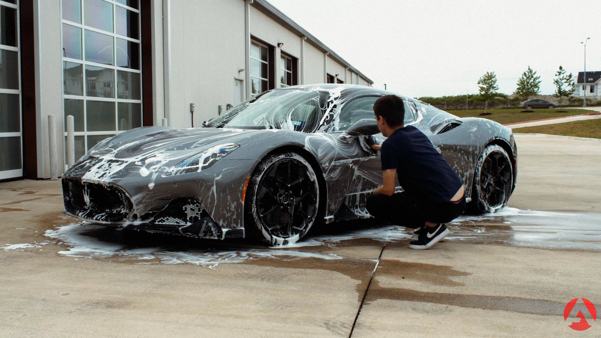 Man washing a gray sports car with black wheels outside a building.