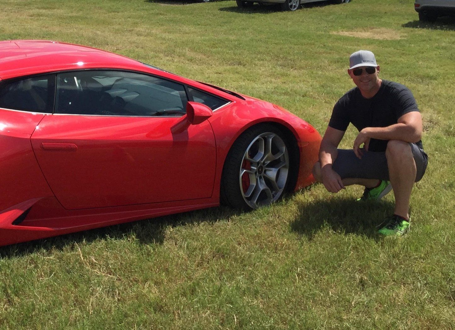 Man squatting next to a red Lamborghini on grassy field. Man is wearing a hat and black t-shirt.