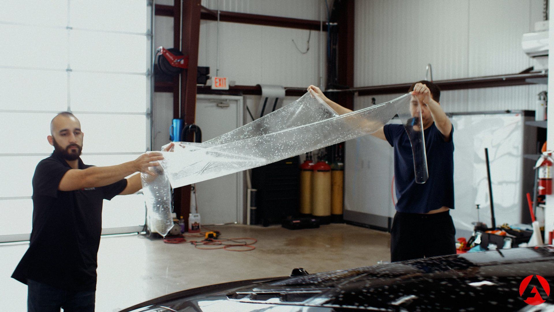 Two men holding up a clear sheet, likely protective film, in a shop; one examines it closely.
