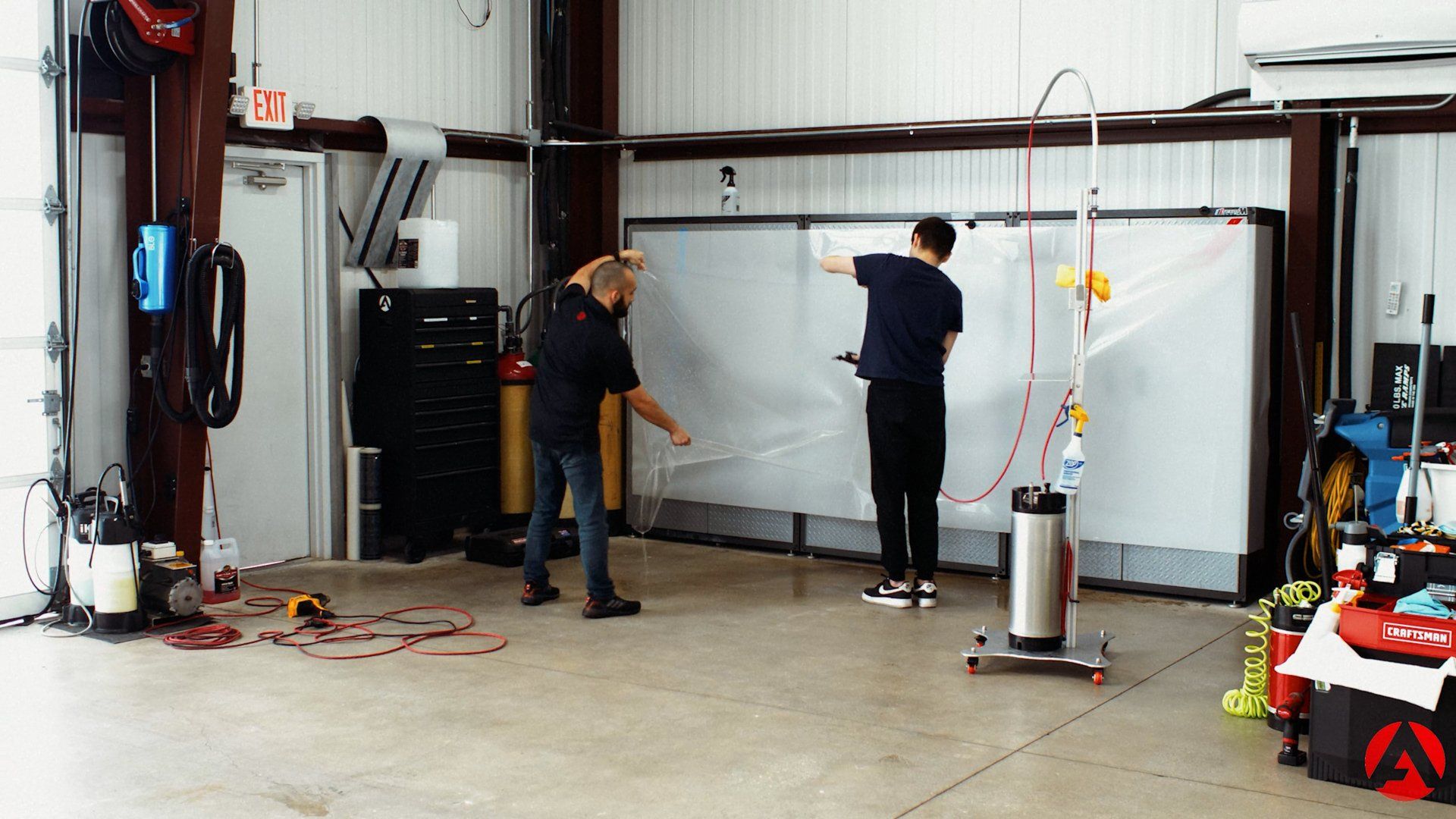 Two men working on a large, clear sheet of material in a garage. One sprays it while the other pulls it.