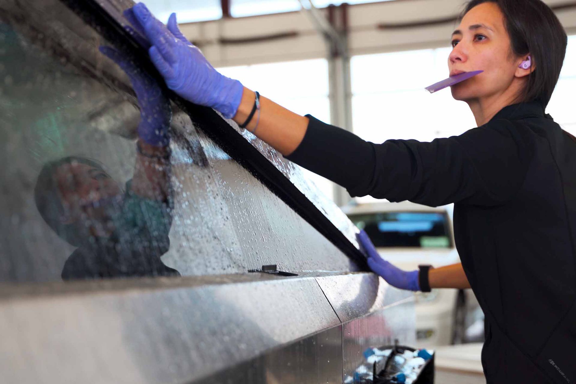 Woman in gloves cleans vehicle window, wearing earplugs, working outdoors.
