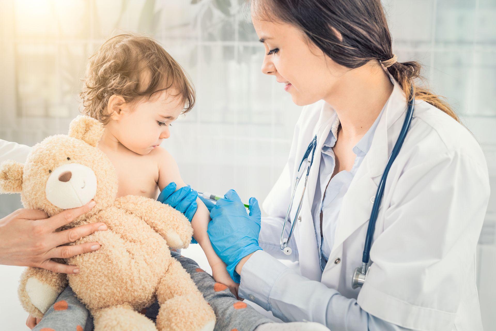 Pediatrician examining a toddler holding a teddy bear in a bright clinic.
