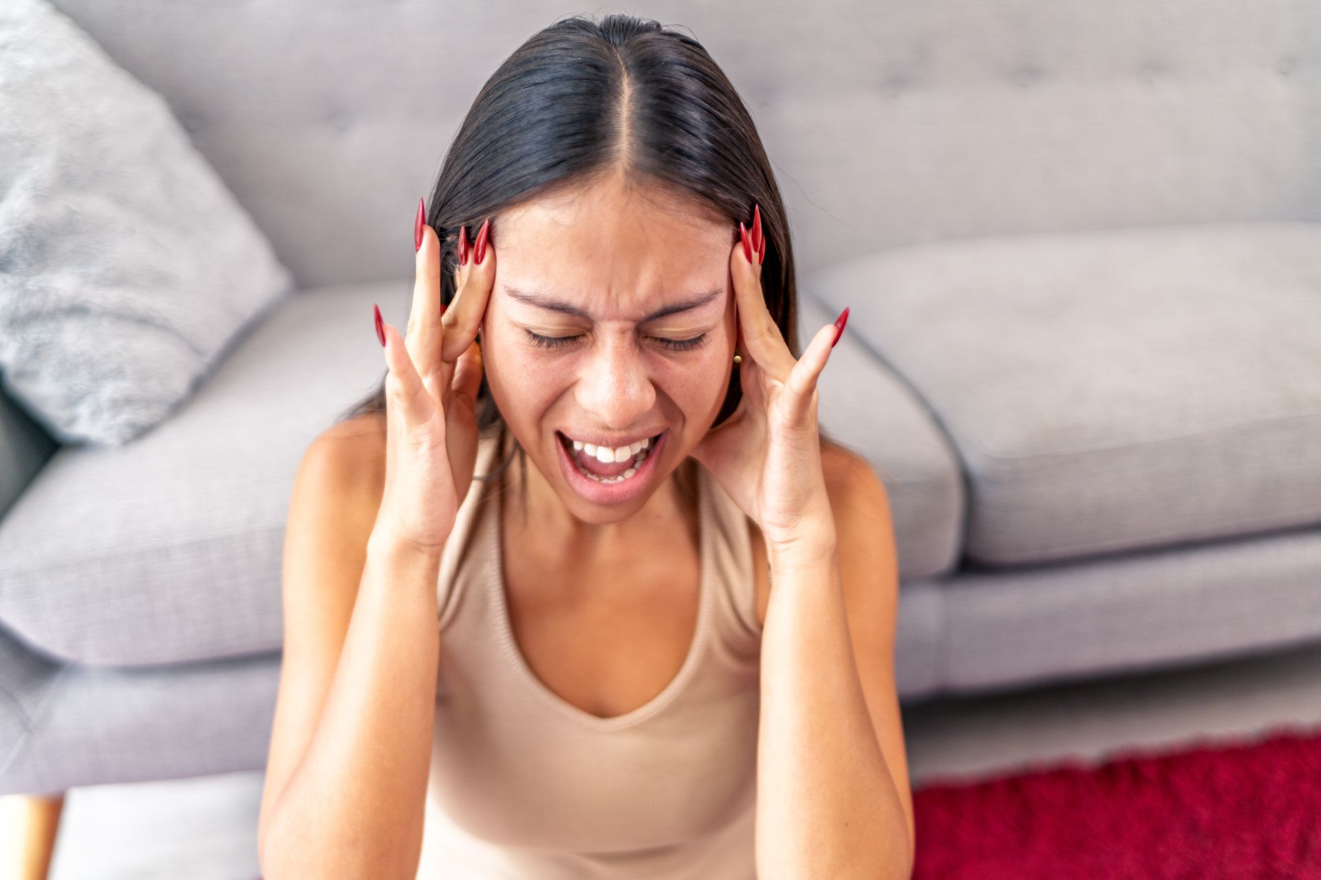 Woman sitting on the floor, holding her head and grimacing in pain in a living room