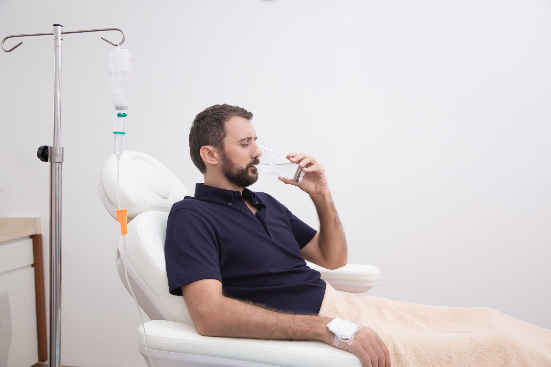 Man reclining in a white chair drinking from a small bottle beside an IV stand