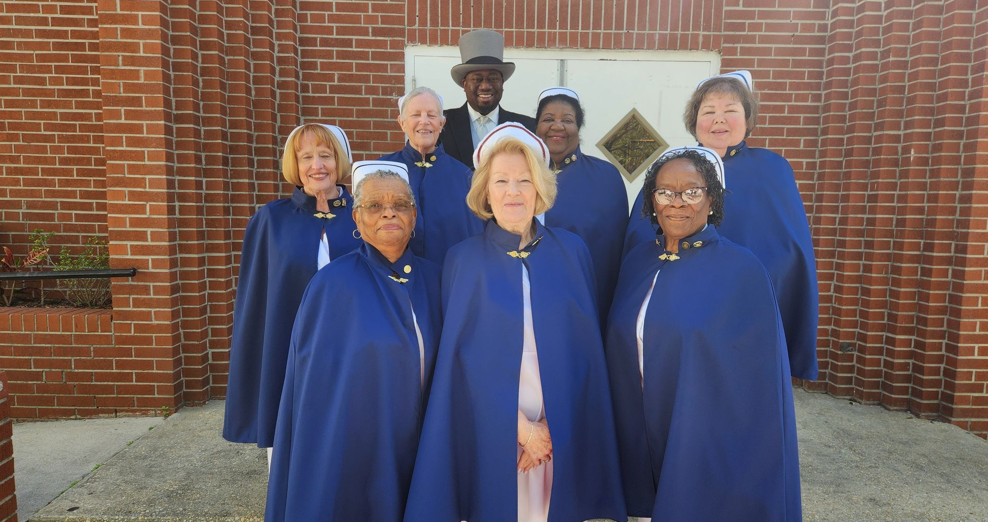 Group of people in blue capes with white trim pose in front of a brick building. One man wears a top hat.