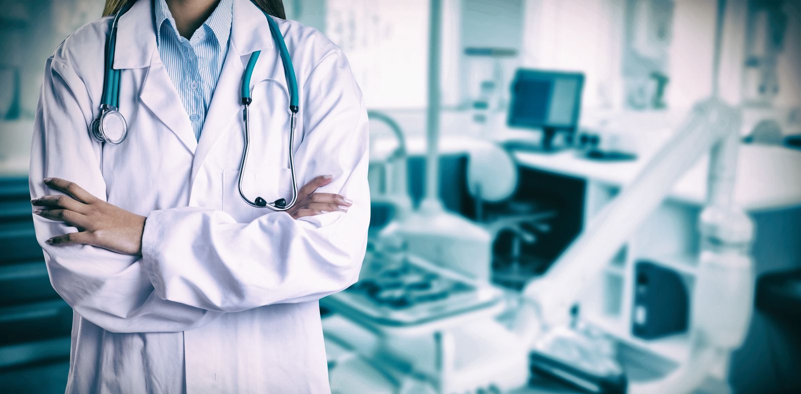 A female doctor with a stethoscope around her neck is standing in a hospital room with her arms crossed.